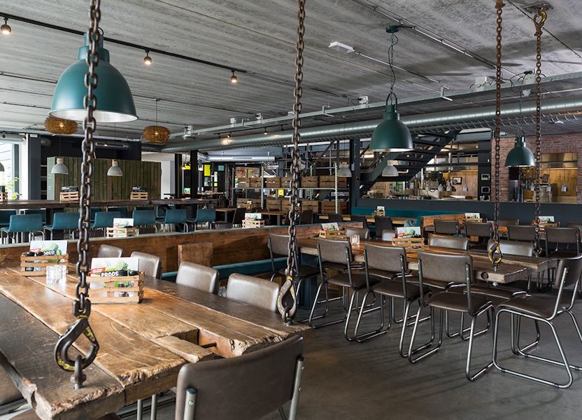 Houten tafels, metalen stoelen en industriële lampen in een ruim restaurant bij Klok'uus, Zeeland; val naar boven.