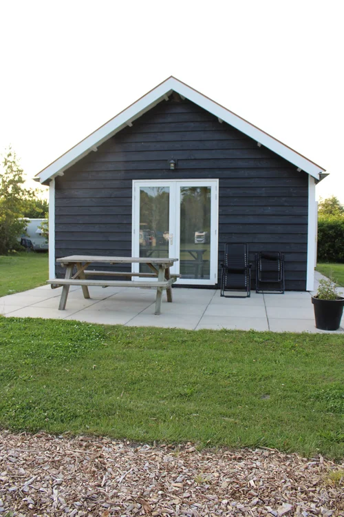 Zwart houten huisje met witte details op Camping De Kolibrie, glazen deuren, picknicktafel, stoelen en planten op terras.