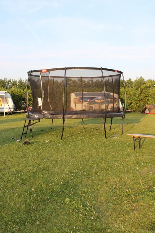 Op Camping De Kolibrie staat een trampoline met veiligheidsnet op gras, omringd door caravans, tenten en bomen onder een heldere lucht.