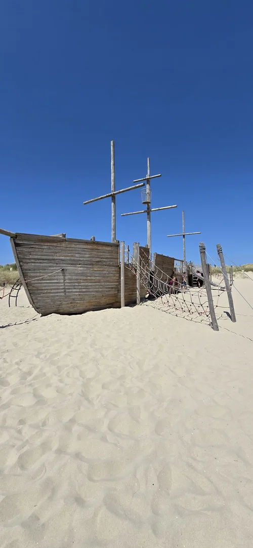 Houten scheepswrak-speeltoestel op het zand bij Camping De Kolibrie, met twee masten en touwen, duinen op de achtergrond.