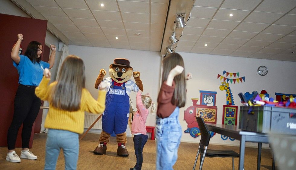 Mascottebeer met drie kinderen en een volwassene in de speelruimte van Hof van Zeeland, iedereen steekt armen omhoog.
