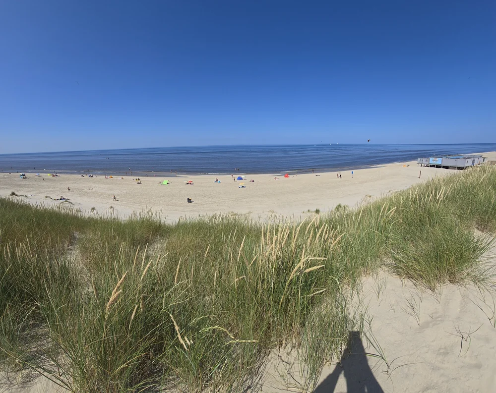 Zandstrand met grasduinen, verspreide mensen en gekleurde parasols langs de kust bij Camping De Kolibrie onder blauwe lucht.