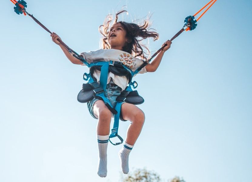 Kind met lang haar in tuigje zweeft aan bungeetrampoline bij uitje Klok'uus, Zeeland, benen bungelend en armen wijd.