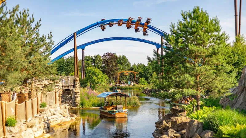 Een bootje vaart over een smalle rivier tussen bomen en rotsen in Toverland, Sevenum; blauwe achtbaan loopt erboven.