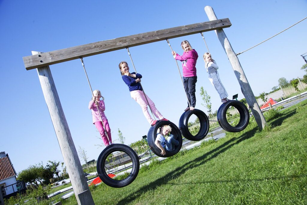 Vijf kinderen spelen op hangbanden van De Jonge Johannes speeltuin, vier staan bovenop en één zit in een band; zonnige dag.