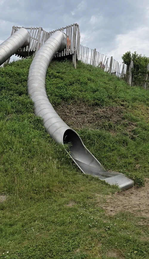 Een lange, bochtige metalen glijbaan komt uit een grasheuvel op Camping De Kolibrie onder een bewolkte lucht.