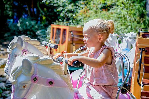 Meisje in roze jurk lacht op witte carrouselpaard buiten bij Avonturenpark Hellendoorn, met zon en groen op de achtergrond.