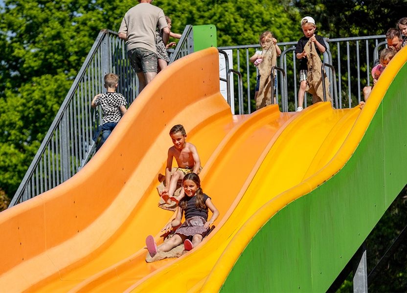Kinderen glijden met jute zakken lachend van de grote gele-oranje glijbaan in Avonturenpark De Bergen, tussen groene bomen.