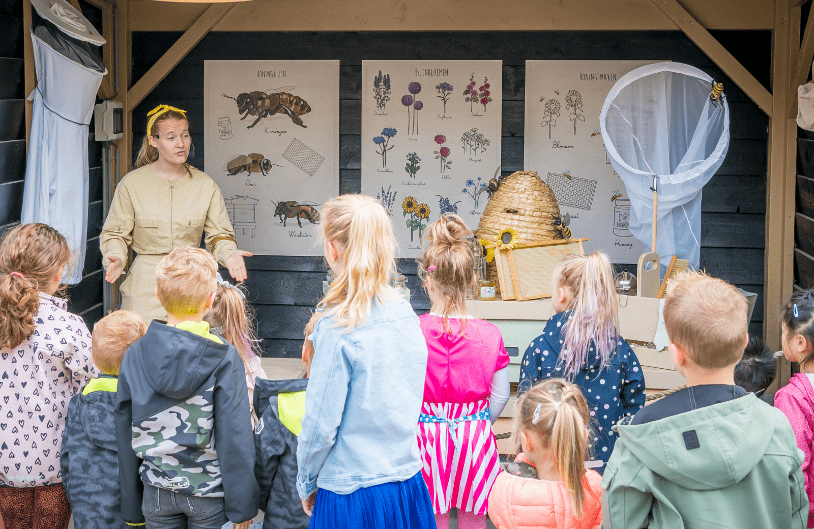 Bij Familie Resort Molenwaard geeft een vrouw in imkerpak buiten les aan kinderen bij bijenposters en imkerspullen.