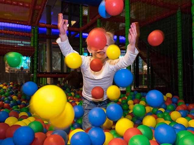 Kind in wit shirt gooit vrolijk rode, gele en blauwe ballen omhoog in kleurrijke ballenbak bij Dinoland Zwolle, uitje.