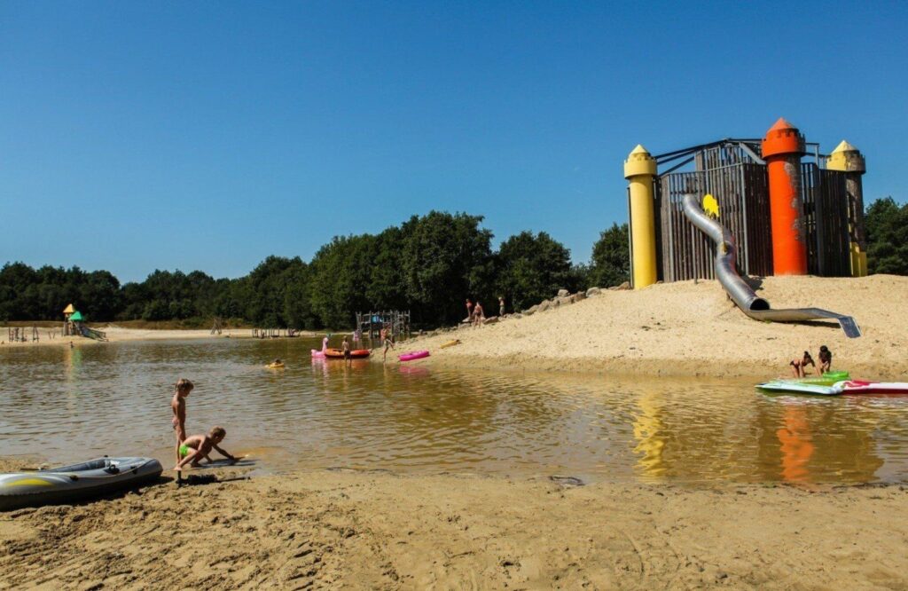 Kinderen spelen met opblaasbare spullen aan een zandstrand bij een kleurrijke speeltuin, omringd door bomen en blauwe lucht.