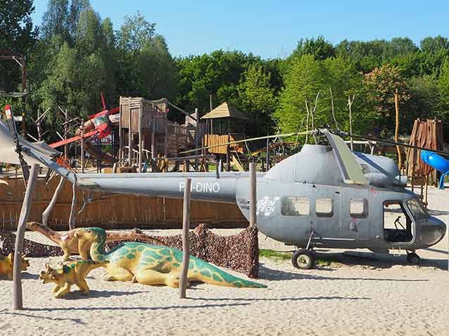 Speeltuin in Dinoland Zwolle met grijze helikopter, dinosaurussen en houten toestellen op zand, omringd door bomen.