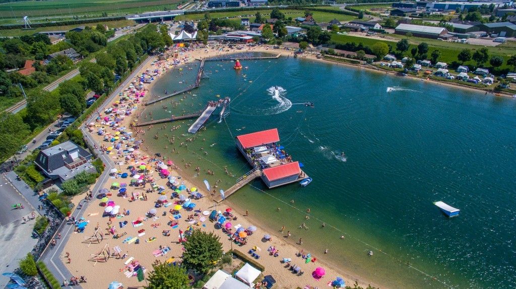 Drukke zandstrand aan meer bij Camping in de Betuwe, Gelderland, met felgekleurde parasols, pier en groene velden.