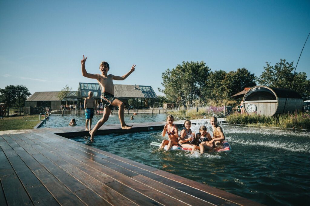 Jongen in zwembroek springt met vredesteken in buitenzwembad terwijl een groep op luchtbedden lacht, vriendelijk en dicht bij zee.