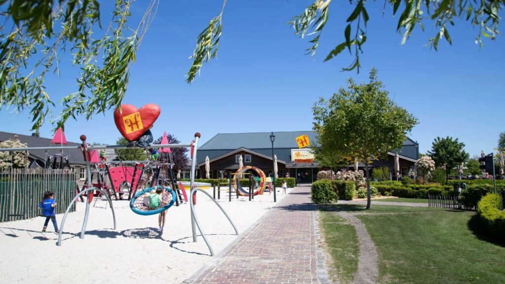 Kinderen spelen in de speeltuin van Hullie Uden met schommels, klimtoestellen en zand onder een blauwe lucht met groen en gebouw.