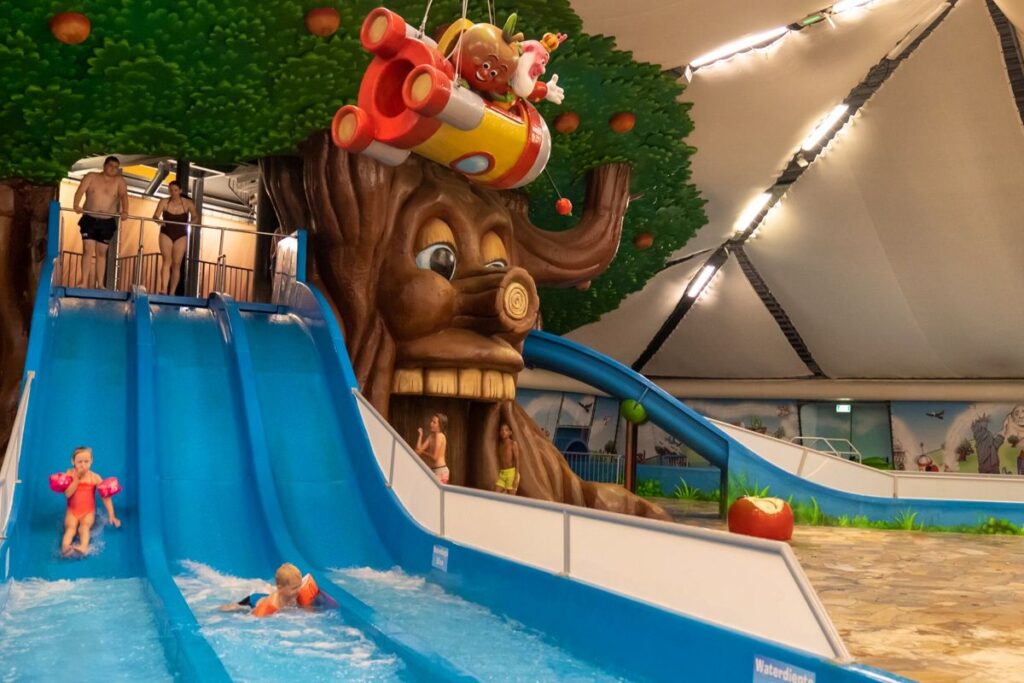 Kinderen spelen op blauwe waterglijbanen naast een boom met gezicht, stripfiguren en rode vliegtuig in een overdekt zwempark.