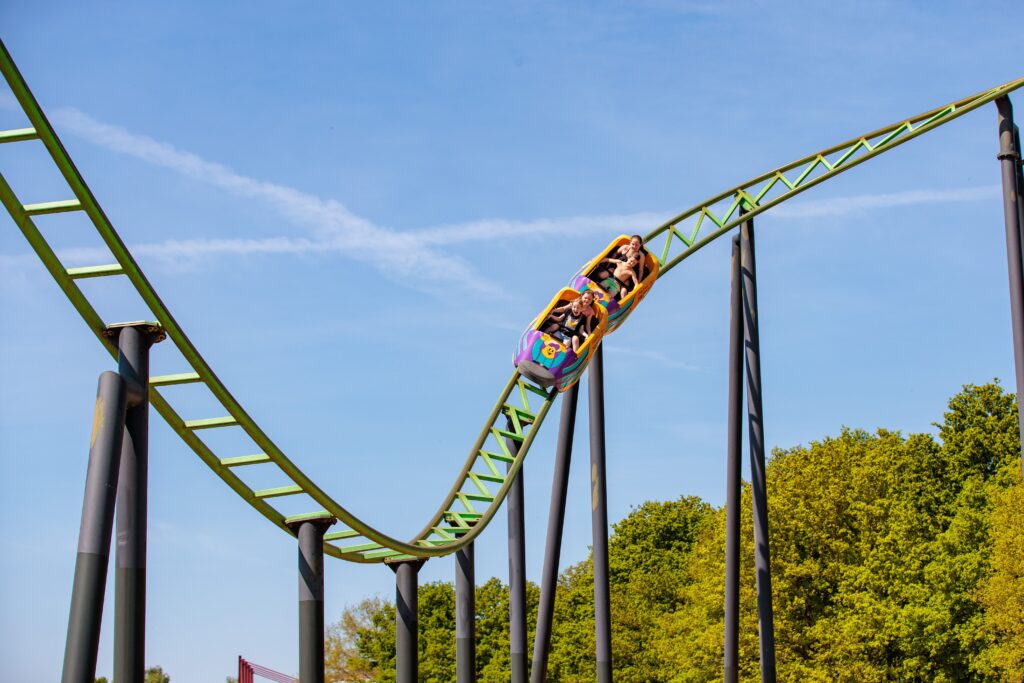 Mensen gaan in een kleurrijke achtbaan met groene rails op BillyBird Hemelrijk omlaag, blauwe lucht en bomen achter ze aan.