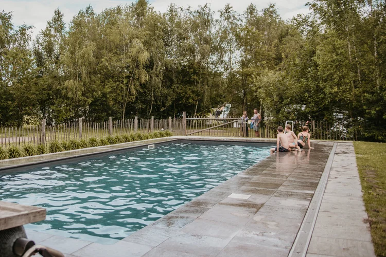 Vriendelijk campingzwembad omgeven door bomen en houten hek, enkele mensen aan de rand en bewolkte lucht.