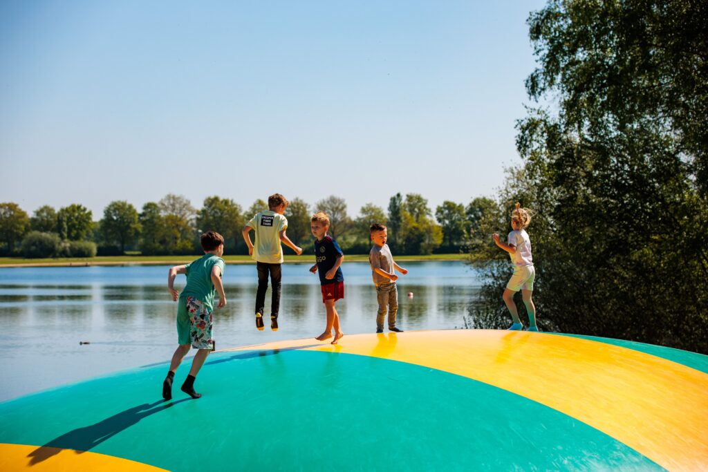 Kinderen spelen en springen op een groot, kleurrijk springkussen bij BillyBird Hemelrijk, met meer, bomen en blauwe lucht.