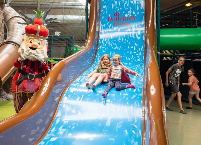Twee jonge meisjes glijden van een grote blauwe glijbaan met een personage in rode kroon; volwassenen kijken lachend toe.