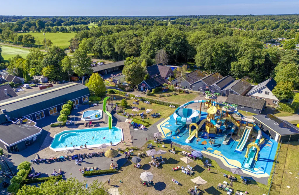 Bovenaanzicht op kindvriendelijk waterpark met kleurrijke glijbanen, zwembaden, ligstoelen en omliggend groen bij mooi weer.