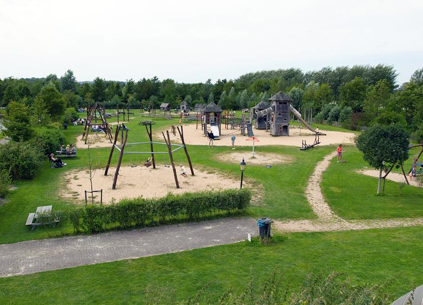 Overzicht van een kindvriendelijke speeltuin met schommels, glijbanen en zand, omringd door gras, bomen en paden.