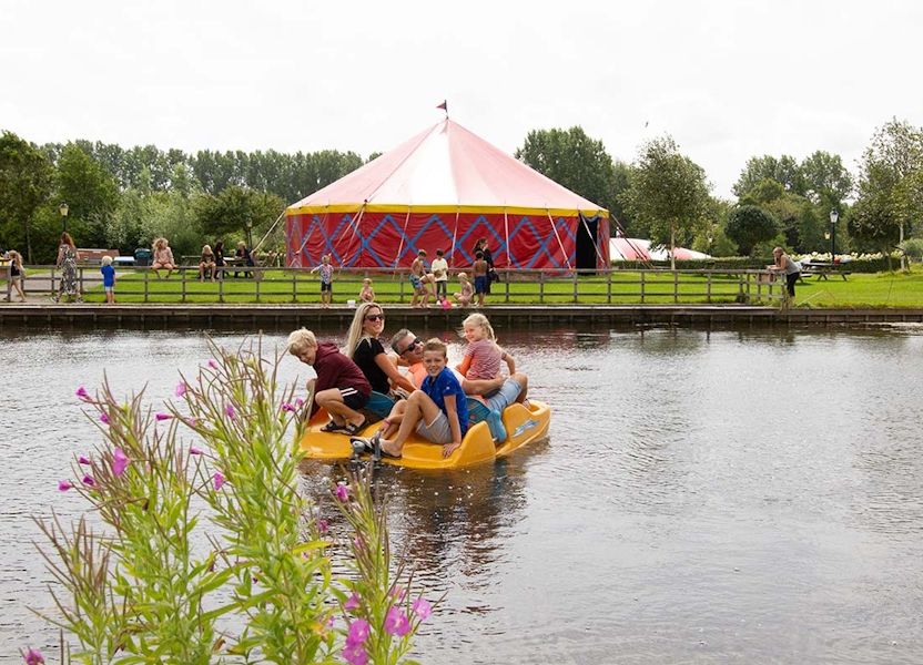 Gezin op gele waterfiets op vijver met roze bloemen, mensen bij rood-geel circustent in groen park, vriendelijk.