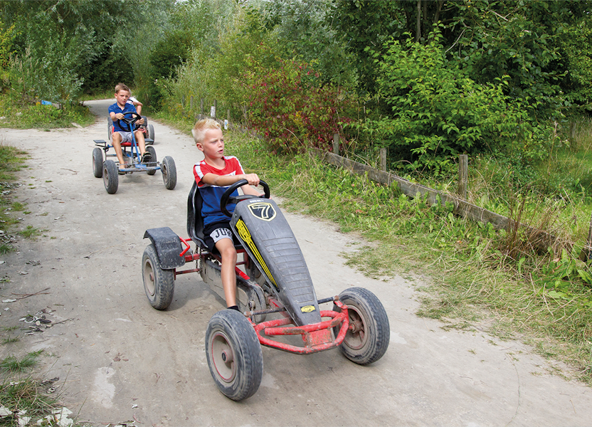 Twee jongens rijden serieus op trapkarts over een bospad, omgeven door groen; de een in rood, de ander in blauw.