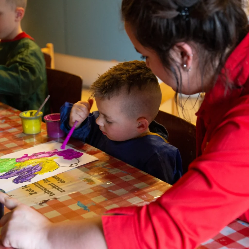 Een kind schildert met paars en groen terwijl een vrouw in rood helpt aan tafel vol knutselspullen bij Hullie Uden, Noord-Brabant.
