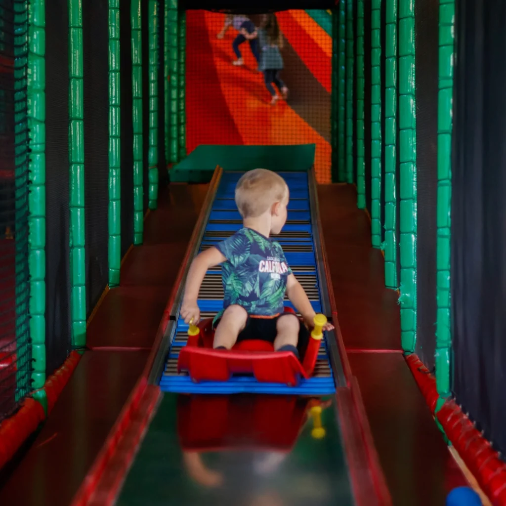 Een jong kind zit op een rode slee klaar om van een kleurrijke glijbaan te gaan bij Hullie Uden, met andere kinderen achter.