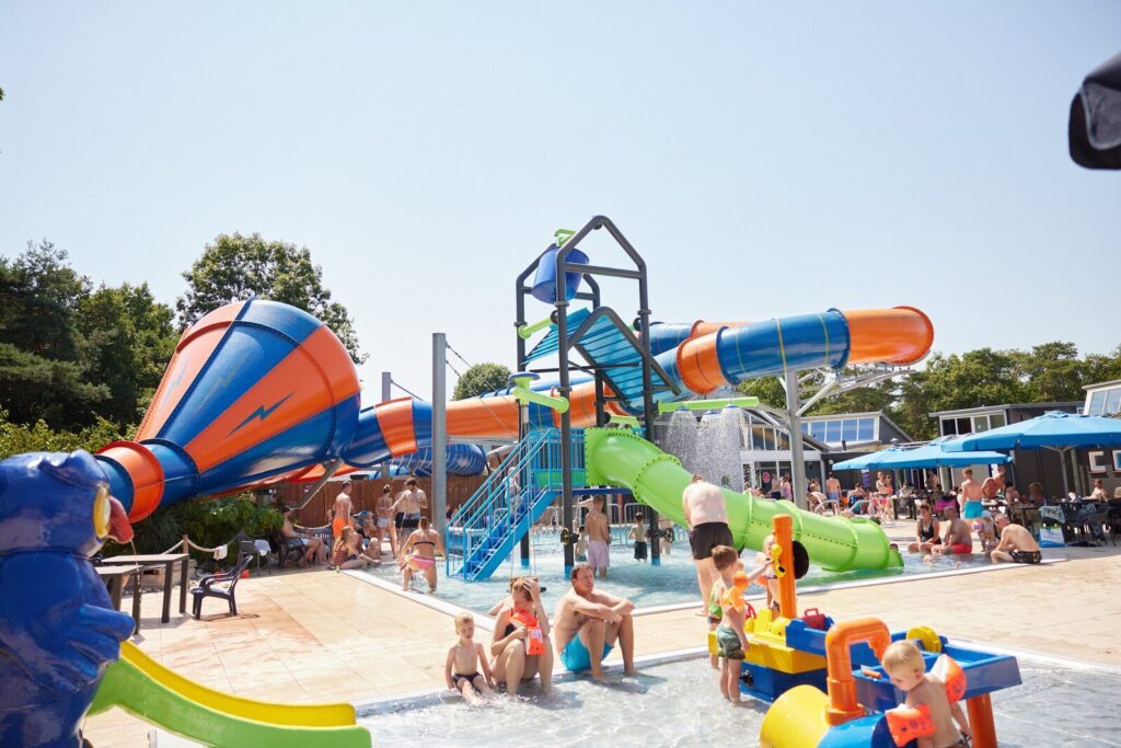 Kinderen en volwassenen spelen bij waterpark met glijbanen en splashzone op kindercamping, Overijssel, onder zonnige lucht.