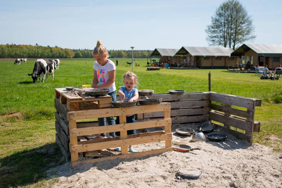 Twee kinderen spelen in een zandkeuken van pallets bij FarmCamps t Looveld, met koeien en tenten op het groene veld.