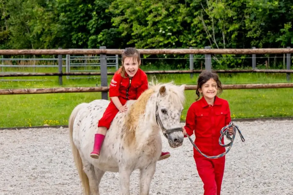 Twee kinderen in rode outfits op FarmCamps De Geele Bosch, één lachend op een witbruine pony, één leidt de pony aan de teugel.