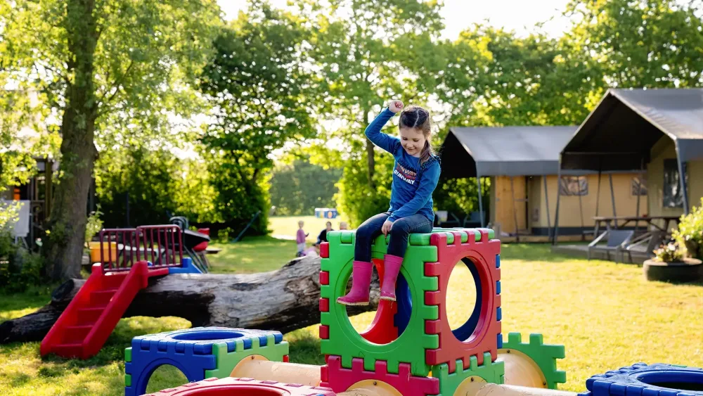 Meisje met roze laarzen en blauwe outfit op kleurrijke speelblokken bij speeltuin FarmCamps Five Star tussen bomen en tenten.