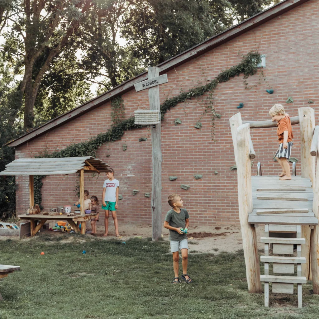 Vier kinderen spelen bij een houten speeltoestel en modderkeuken, met klimstenen tegen een muur op de achtergrond.