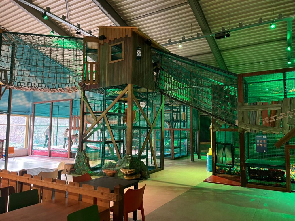 Grote indoorspeeltuin bij Landal Coldenhove met houten klimmen, touwenbruggen, glijbanen en tafels met gekleurde stoelen.