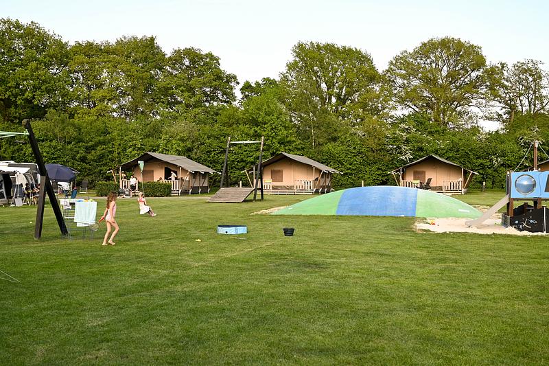 Kind loopt op het gras bij Camping Dal van de Mosbeek met tenten, speeltuin en springkussen; bomen onder heldere lucht.